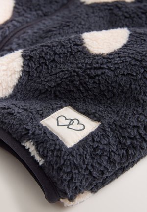Tejido de forro polar azul marino suave con lunares crema y una etiqueta tejida que presenta corazones entrelazados. Textura suave, cierre con cremallera.