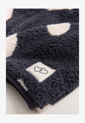 Fuzzy marineblå fleece-stof med crème farvede prikker og et vævet mærke med sammenflettede hjerter. Blød tekstur, lynlås lukning.