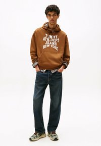 Sweatshirt à capuche marron avec texte blanc, jean en denim bleu et baskets grises avec des accents noirs et rouges. Le modèle pose les mains dans les poches.