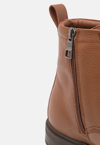 Bottine en cuir brun avec fermeture éclair sur le côté, surface texturée et un petit tirant pour un enfilage facile. Semelle en caoutchouc durable visible.