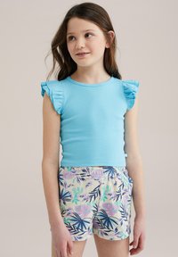 Vaaleansininen ribbitoppi, jossa on röyhelökoristeet olkapäillä, yhdistettynä kermaisiin shortseihin, joissa on värikkäitä trooppisia lehtikuoseja.