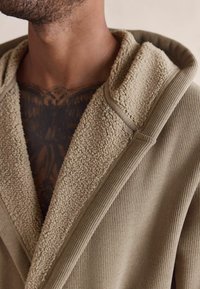 Beige kapuutsiga ese, millel on tekstuuriga, pehme fliisvooder kaeluses ja soonikmaterjal. Suurendatud vaade toob esile disainidetailid.