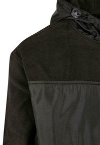 Svart jakke med teksturert fleece øvre del og glatt nedre del, med snorhette og kontrasterende stoffstrukturer.