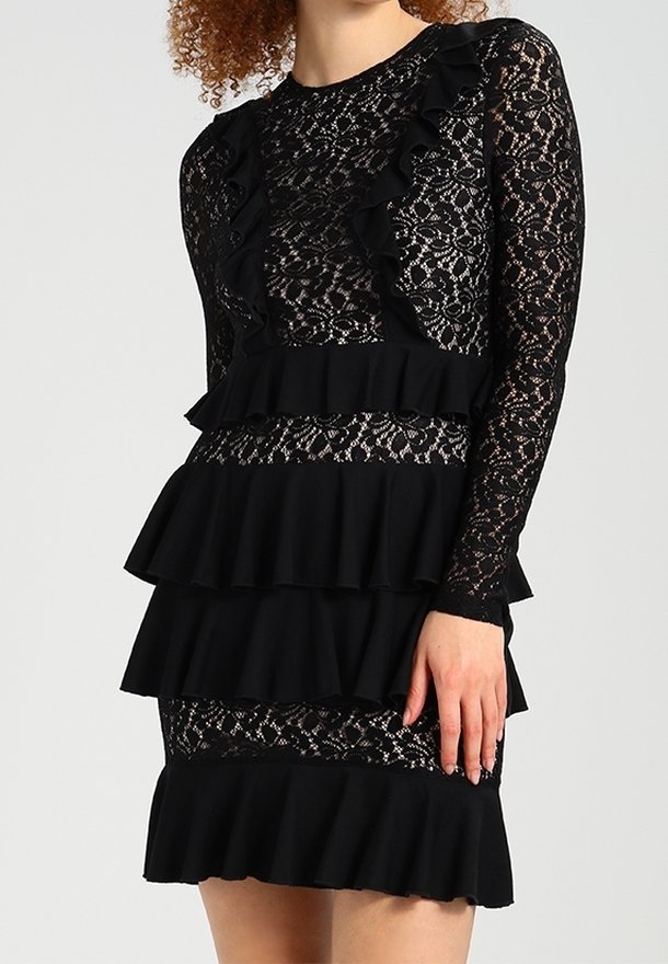 Femme portant une robe en dentelle noire avec des manches longues et des détails en volants à plusieurs niveaux au niveau de la poitrine, de la taille et de l'ourlet.