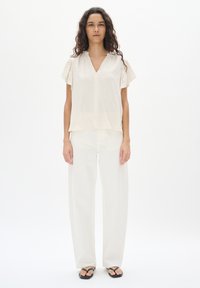 Top beige chiaro a maniche corte con scollatura a V, realizzato in tessuto texturizzato, abbinato a pantaloni bianchi a gamba larga e infradito neri. Look semplice e rilassato.