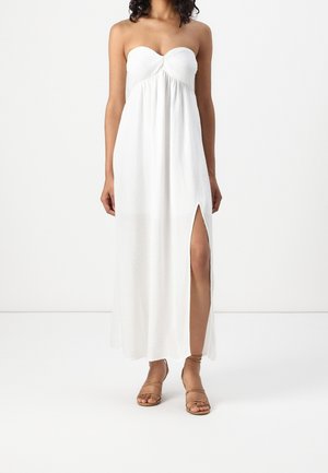 Kobieta ubrana w białą suknię maxi bez ramiączek z rozcięciem po boku oraz beżowe sandały na paski, stojąca na jednolitym białym tle.