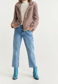 Femme portant une veste en fausse fourrure marron clair, un col roulé blanc, un jean court bleu et des bottines bleues, debout les mains dans les poches.