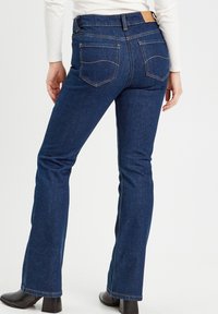 Dunkelblaue, ausgestellt geschnittene Jeans aus Denim mit hoher Taille, betonten Gesäßtaschen und einem hellbraunen Lederetikett am hinteren Bund. Glatte Textur.