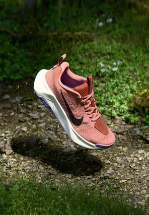 Roze Nike hardloopschoen met zwarte swoosh zwevend boven een zandpad met gras in het zonlicht.