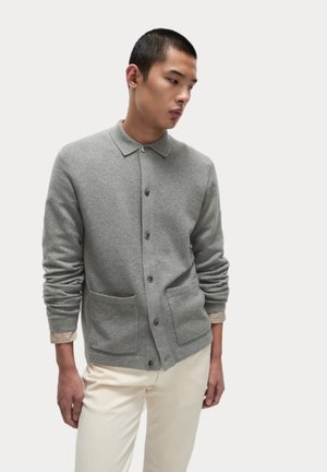 Cárdigan gris hecho de material suave, con un cierre de botones en la parte delantera, dos bolsillos frontales y un cuello con solapa. Lleva una camiseta de rayas debajo.