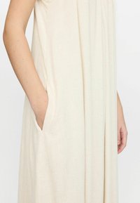 Cremefarbiges Kleid mit entspannter Silhouette, ausgestattet mit Seitentaschen und weichem, strukturiertem Stoff. Die Silhouette ist locker und fällt sanft.