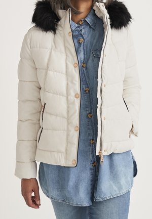 Mujer con chaqueta beige acolchada con capucha de pelo sintético negro, sobre camisa vaquera azul con botones y vaqueros, manos relajadas a los lados.