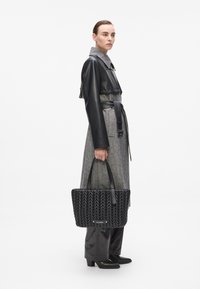 Sac fourre-tout noir tissé avec un design structuré, poignées en cuir lisse et petite plaque logo en métal. Modèle portant un manteau gris et des bottes.
