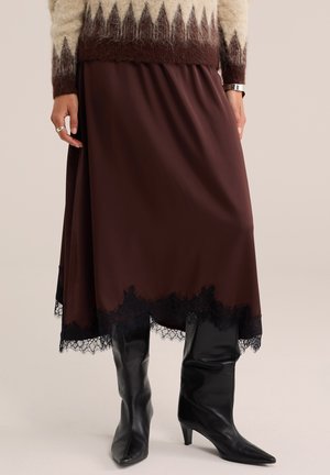 Personne portant une jupe marron avec bordure en dentelle noire, des bottes noires montantes à talons et un pull beige et marron à motif duveteux.