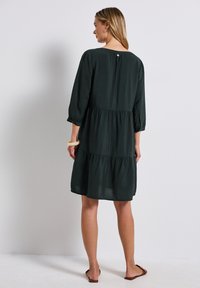 Musta vihreä, kerroksellinen mekko pitkillä hihoilla, pehmeä rakenne ja takana nappidetali. Suunniteltu rennoksi ja polven pituiksi.