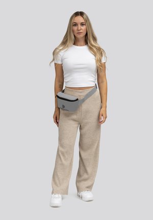 Grijze heuptas met een ritssluiting en logo, gedragen over een wit crop top en beige brede broek. Witte sneakers maken de outfit compleet.
