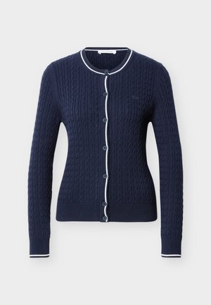 Cardigan bleumarin cu model tricotat în stil cablu, cu bordură albă pe guler, banda de nasturi și manșete, având nasturi în față și mâneci lungi.