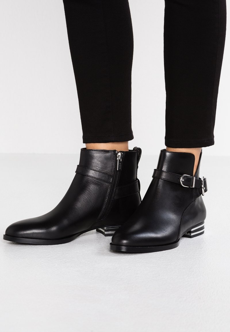 Jambes inférieures vêtues d'un pantalon noir slim et de bottines noires à fermetures éclair latérales et sangles à boucle, debout sur un sol de couleur claire.