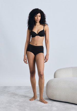 Reggiseno nero e slip a vita alta, entrambi realizzati in tessuto liscio. Modella in piedi scalza su una superficie grigia chiara e testurizzata. Sfondo minimalista.