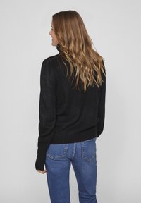 Svart ribbad tröja med hög krage, ett par blåjeans och långt vågigt hår. Synlig texturerad stickning och figurnära midjedesign.