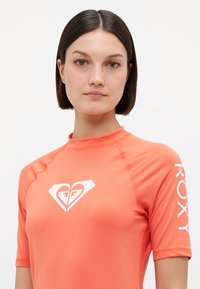 Kortermet korallfarget rashguard laget av elastisk materiale med en hvit hjerte-logo på forsiden og "ROXY" trykt vertikalt på ermet.