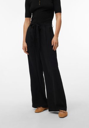 Femme portant un pantalon large noir taille haute avec ceinture nouée, un haut noir à manches courtes et des ballerines beiges.