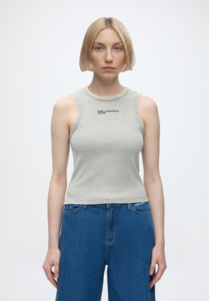 Jeune femme aux cheveux blonds courts portant un haut gris clair sans manches avec le texte « Karl Lagerfeld Jeans » et un jean en denim bleu.