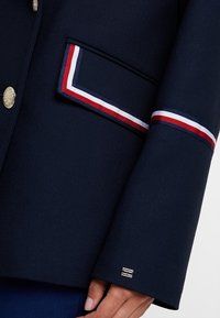 Manche et poche d'un blazer marine avec bordure rouge, blanche et bleue, boutons décoratifs argentés et petit logo métallique près du poignet.