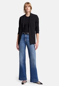 Schwarzer Blazer über einem schwarzen Oberteil, kombiniert mit weit geschnittenen, verblassten blauen Jeans und gemusterten Ballerinas. Merkmale sind der taillierte Schnitt und die Umschlagärmel.