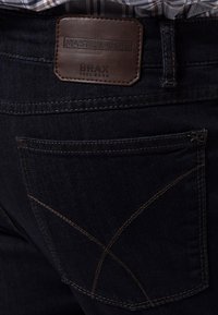 Vaqueros de mezclilla oscura con una etiqueta de cuero etiquetada "BRAX FEEL GOOD" y un diseño de costura estampada en el bolsillo trasero.