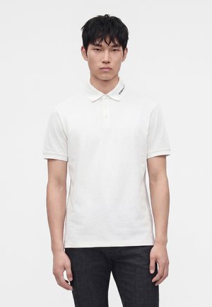 Jeune homme portant un polo blanc uni à trois boutons et un pantalon noir, debout devant un fond blanc.