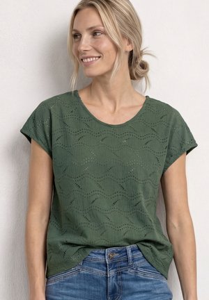 Kvinde med blondt hår i en lav knold iført en grøn øjlet kortærmet bluse og blå jeans, smilende og kigger mod venstre.