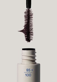 Maskaraharja nostamassa tummaa maskaraa valkoisesta putkesta, jossa lukee "85+ / 100", vaaleaa taustaa vasten.