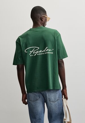 Homme portant un t-shirt vert avec du texte blanc "Pegador All Rights Reserved" dans le dos, jean bleu, lunettes de soleil, et tenant un sac fourre-tout beige.
