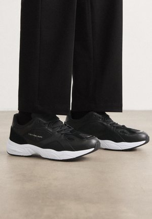 Baskets Calvin Klein Jeans en noir et blanc portées avec un pantalon noir, debout sur une surface lisse de couleur brun clair.