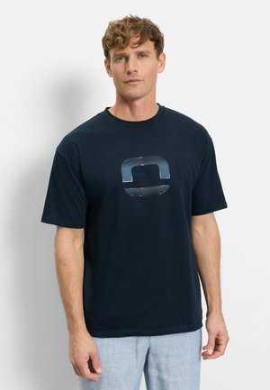 Mann mit hellbraunen lockigen Haaren, der ein marineblaues T-Shirt mit einem reflektierenden geometrischen Logo und hellgraue Hose trägt, steht vor einer weißen Wand.