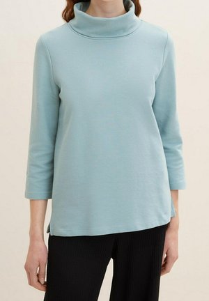 Kvinde iført lyseblå turtleneck med trekvartlange ærmer og sorte ribstrikkede bukser, stående mod en ensfarvet baggrund.