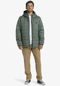 Quilted green jacket with a hood, front zipper, and logo patch. Worn over a white shirt and tan pants with black sneakers.