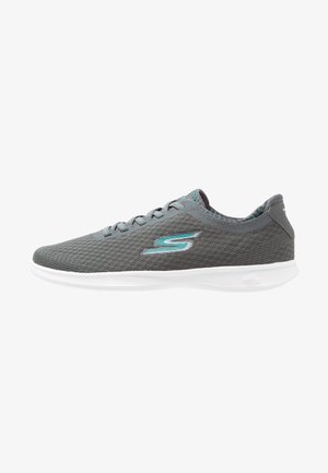 Zapatilla deportiva de malla gris con detalle en verde azulado, logo "S", suela blanca y diseño con cordones, vista desde el lado exterior.