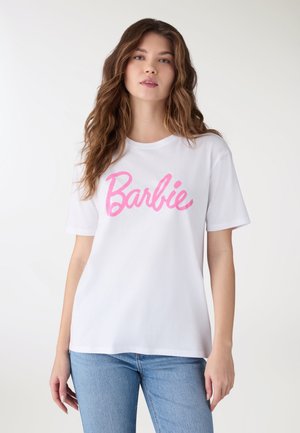 Giovane donna con capelli castani mossi che indossa una maglietta bianca con la scritta "Barbie" in rosa e jeans azzurri chiari su uno sfondo semplice.