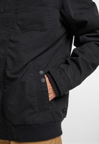 Chaqueta exterior negra con cierre de cremallera, puños acanalados, bolsillos laterales con detalles de botón y una textura de tela suave y mate.