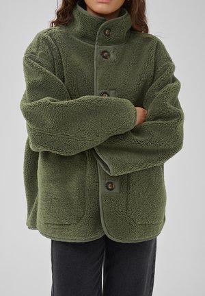 Personne portant une veste polaire verte oversize avec de gros boutons et un col montant, les bras croisés, visible des épaules aux hanches.