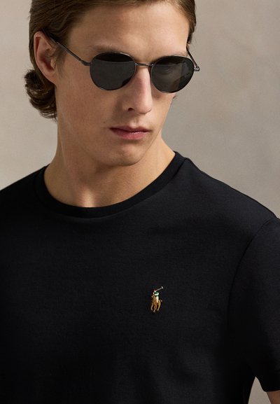 Junger Mann trägt runde schwarze Sonnenbrille und ein schwarzes T-Shirt mit kleinem besticktem Polo-Spieler-Logo auf der Brust.
