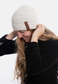 Kremowa dziana czapka z prążkowanym mankietem i brązową skórzaną łatką. Noszona przez osobę z długimi włosami, w połączeniu z czarną kurtką z korduroy.