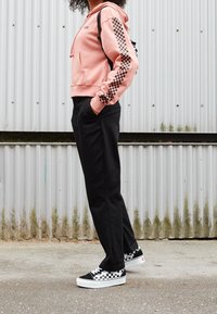 Persona che indossa un felpa rosa con striscia a scacchi nera, pantaloni neri e sneakers a scacchi bianchi e neri, in piedi contro un muro ondulato.