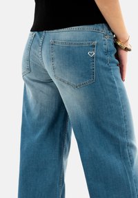 Jean en denim bleu à jambe large avec une finition texturée, comportant deux poches arrière et un détail de couture en forme de cœur sur la poche droite.