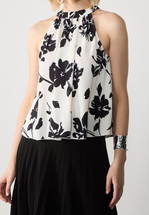 Vrouw draagt een mouwloos wit haltertopje met zwarte bloemenprint en een brede zilveren manchetarmband aan haar rechterpols.