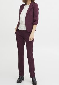 Blazer bordeaux et pantalon assorti avec un chemisier blanc à volants. Le blazer est doté d'un seul bouton et de manches retroussées ; le pantalon est cintré. Chaussures à talons noires.