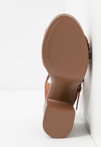 Semelle d'une sandale en cuir marron avec semelle extérieure en caoutchouc texturé, talon carré et bride ajustable à boucle, sur fond blanc.