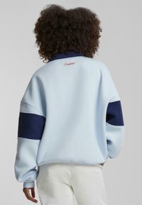 Lyseblå sweatshirt med marineblå detaljer, ribbede mansjetter og høy krage. Har "Forbudt" brodert i rødt på ryggen.